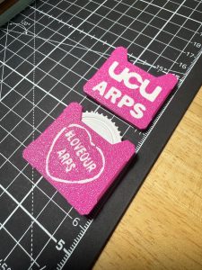 A photo of two pink and white 3D printed fidget toys next to each other showing both sides of the design; one side with the words UCU ARPS on one side and a "love our ARPS" lovehearth logo on the other