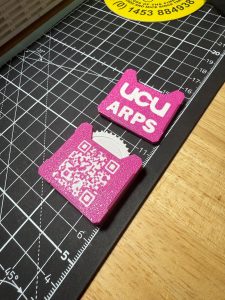 A photo of two pink and white 3D printed fidget toys next to each other showing both sides of the design; one side with the words UCU ARPS on one side and a QR on the other
